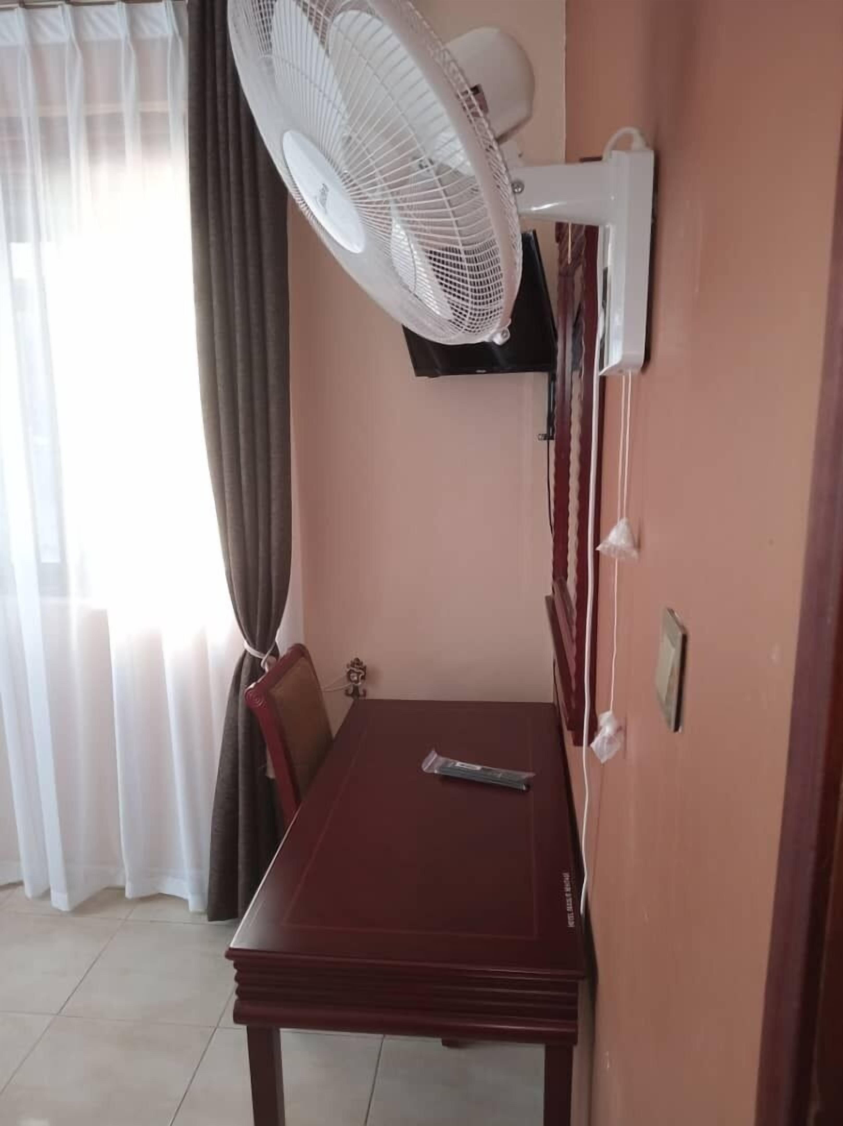 Quarto com uma mesa de jantar, televisão presa à parede, ventoinha no teto e janela com cortinas.