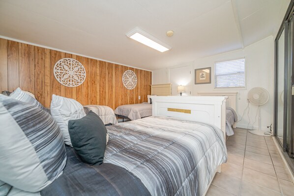 A bedroom with a large bed, two smaller beds, wooden paneling on the wall, a bedside table with a lamp, and a window with blinds. - Secluded coastal retreat (Aransas Pass)