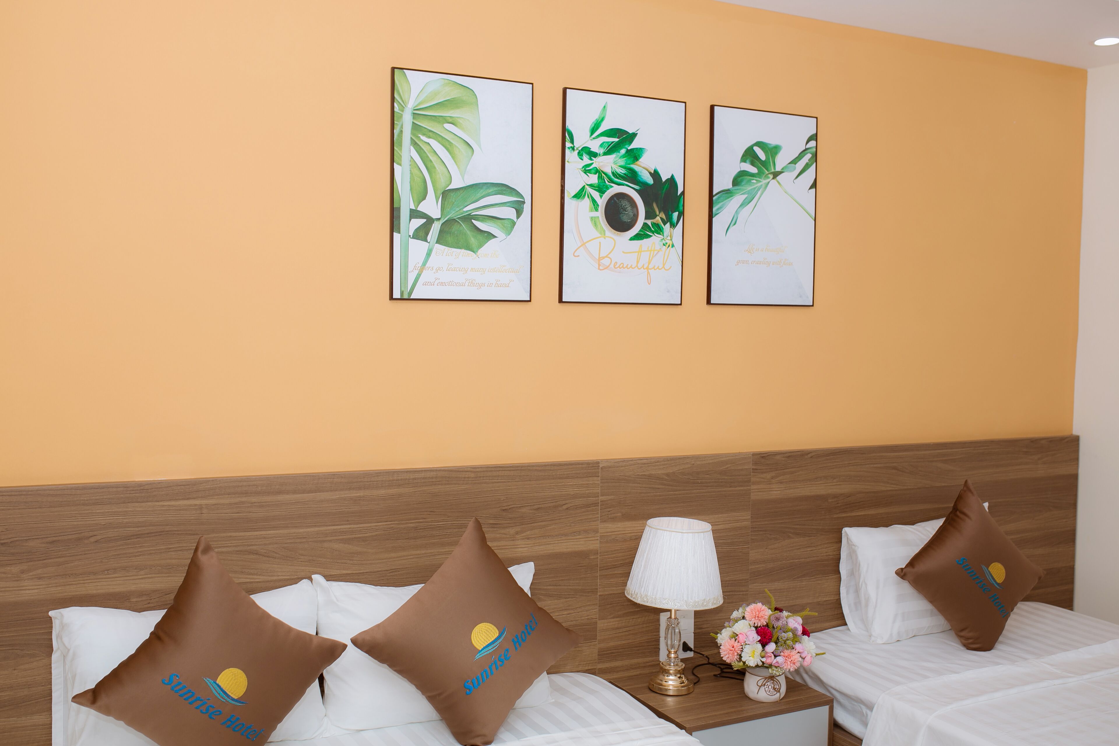 A hotel room with two beds, a nightstand with a lamp, a vase with flowers, and three framed botanical prints on the wall.