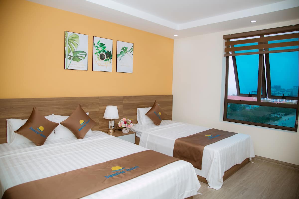 Quarto de hotel com duas camas, cabeceira de madeira, uma janela ampla com vista para a cidade e três quadros com gravuras botânicas na parede.