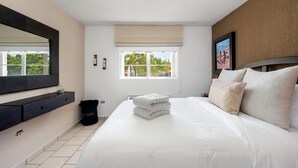 A hotel room with a large bed, a window with a view of palm trees, a black trash bin, and a black wall-mounted shelf.