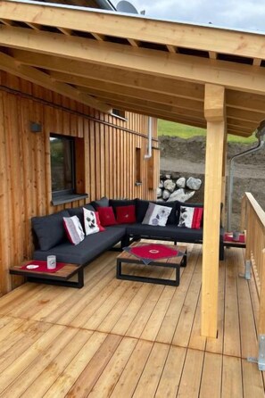 Une terrasse en bois avec un canapé noir et deux tables basses, une fenêtre et un mur de pierre en arrière-plan.