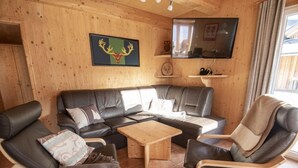 A cozy living room with a sectional sofa, two armchairs, and a coffee table. A television is mounted on the wall, and there is a wooden shelf with decorative items. - Premium #14 mit IR-Sauna by Interhome (Hohentauern)