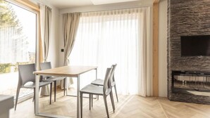 A modern living room with a wooden table and chairs, a stone fireplace, and large windows with curtains.