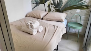 Una cama bien hecha con cubrecama y almohadas color beige, una silla verde y una planta grande al fondo.