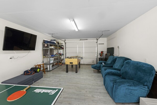 A living room with a foosball table, a sofa, a ping pong table, and a TV mounted on the wall.