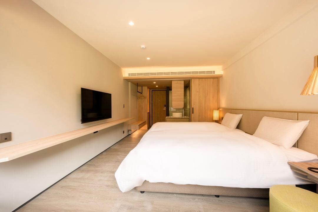 A modern hotel room with a large bed, a mounted TV, a bedside table, and a built-in bench along the wall.