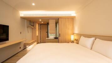 A modern hotel room with a large bed, a TV on the left, a wooden wardrobe, and a bathroom visible through an open door.