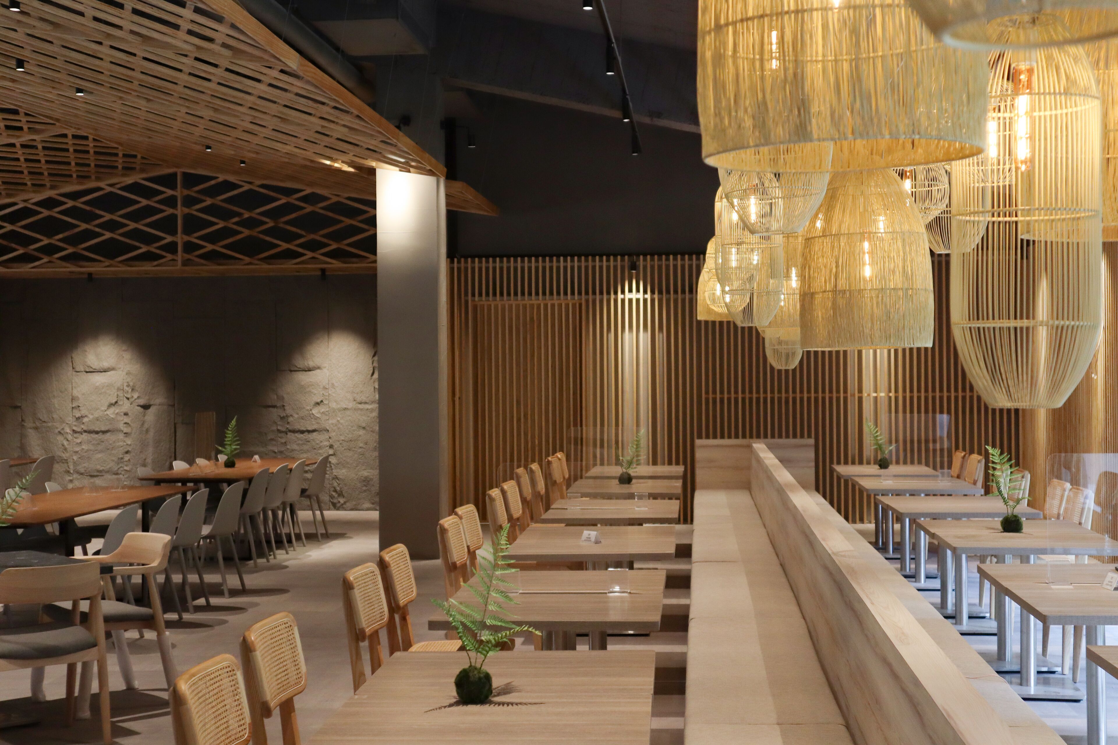 A modern dining area with long tables and chairs, wooden paneling, and hanging light fixtures.