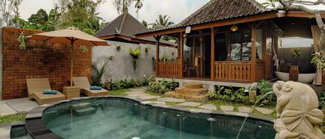 A swimming pool with a stone edge, a wooden deck with lounge chairs, and a covered seating area with a tiled roof.