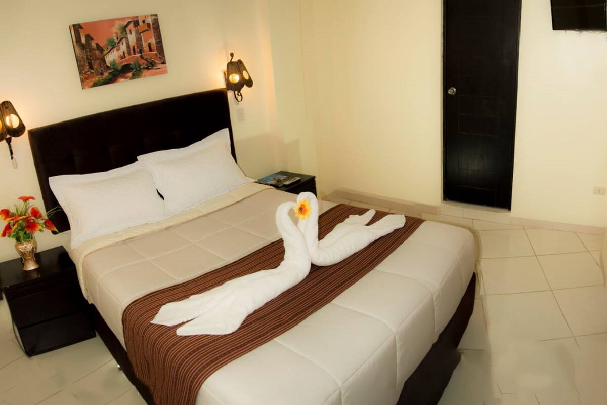 Una habitación de hotel con cama, mesita de noche, lámpara, un cuadro en la pared y una puerta.