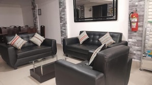 Ein modernes Wohnzimmer mit zwei schwarzen Ledersofas, einem Glastisch und gemusterten Zierkissen. Ein Feuerlöscher ist rechts an der Wand angebracht.