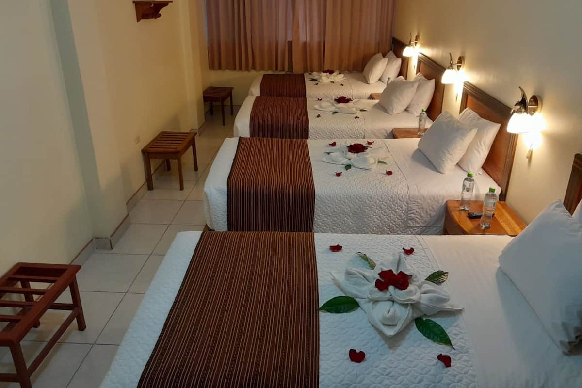 Habitación de hotel con tres camas, cada una con cabecero de madera y ropa de cama blanca. Hay mesitas de noche con lámparas, un taburete pequeño y una decoración de toallas blancas y rosas rojas sobre las camas.