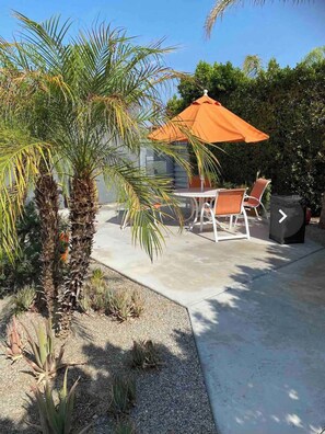 Relax outdoors with a table and two chairs under a spacious umbrella, surrounded by lush greenery.