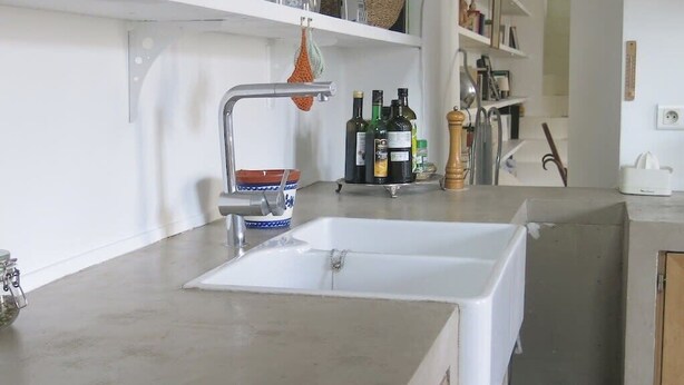 Una cocina con un lavamanos blanco, estantes con vajilla y un florero con flores.