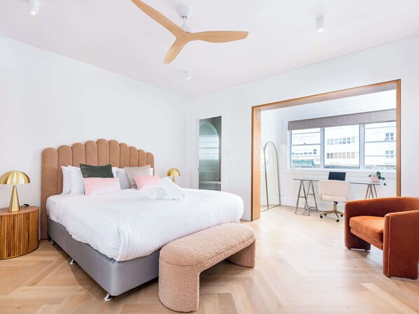 Ein modernes Schlafzimmer mit einem großen Bett, einem Hängeventilator aus Holz, einem Schreibtisch und Blick auf Gebäude durch ein Fenster.