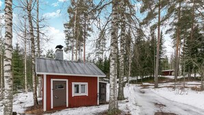 Eine kleine Holzhütte mit einem Metalldach und roter Fassade, umgeben von hohen Bäumen und schneebedecktem Boden.