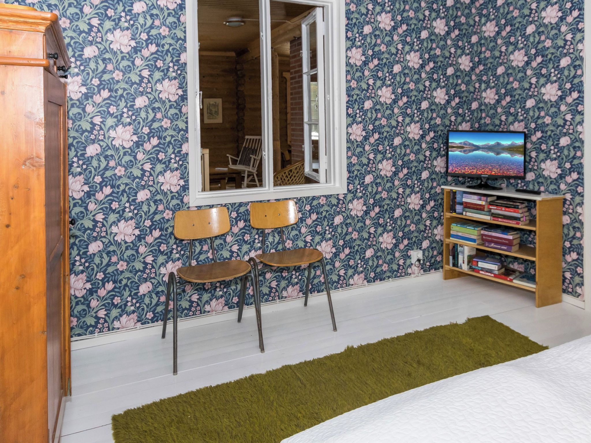 A room with floral wallpaper, a television, a bookshelf, two chairs, and a wooden cabinet.