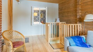 Ein gemütliches Zimmer mit Holzwänden und -decke, einer Holzreling, einem Korbsessel, einer Couch und einem Fenster mit Aussicht.