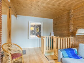 Una habitación con techo de madera, una ventana con papel tapiz floral, una cuna de madera, una silla de mimbre y un sofá con una manta azul.