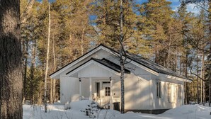 A white cabin with a gabled roof surrounded by snow-covered ground and tall trees.