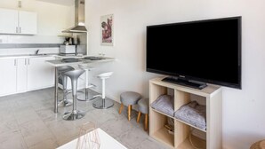 Et moderne køkken med hvide skabe, en rustfri stål emhætte og et barområde med barstole. Et fladskærms-TV er monteret på væggen over en træhylde med puder.