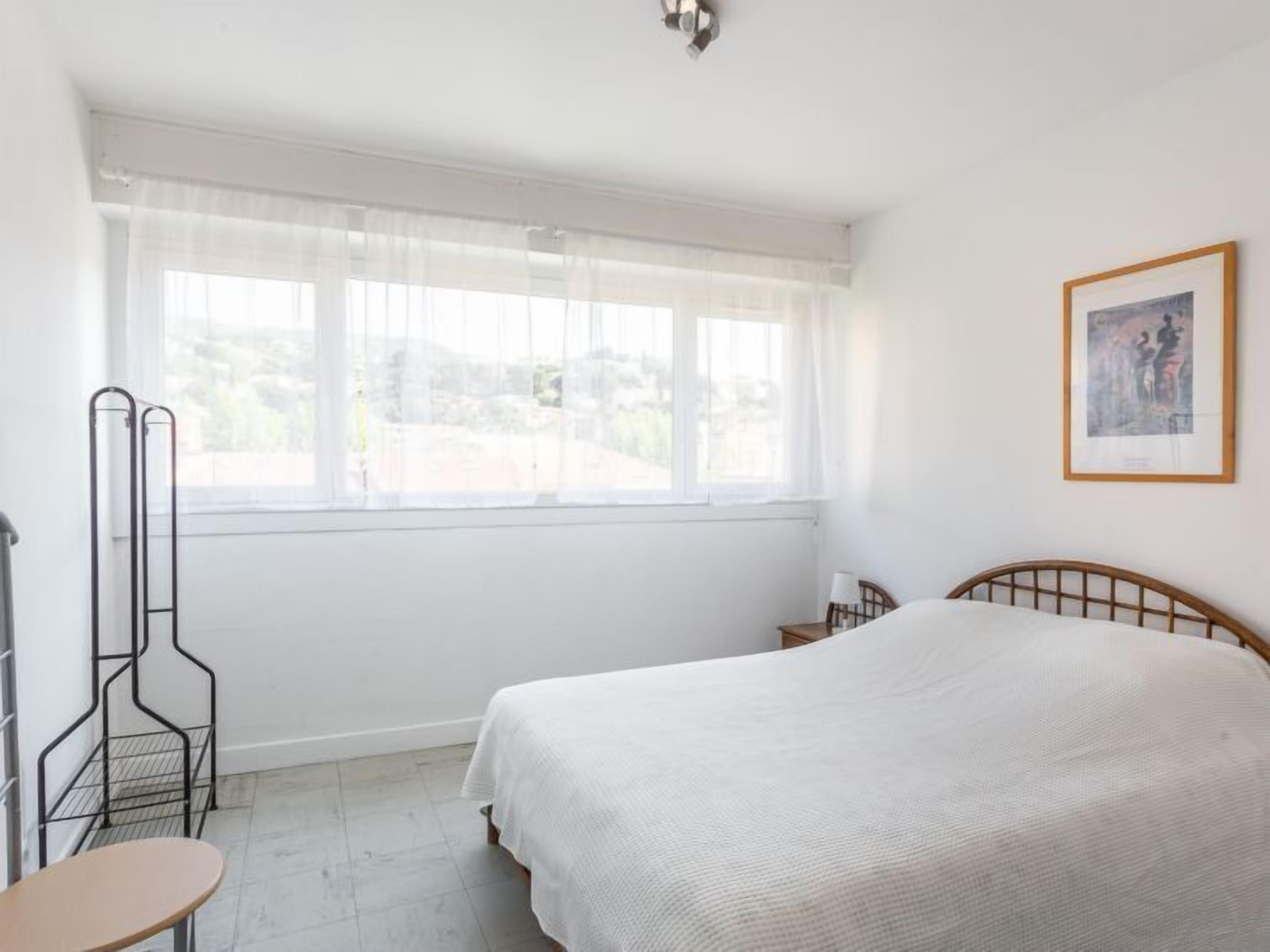 A bedroom with a bed, a window with sheer curtains, a framed picture on the wall, and a metal rack against the wall.