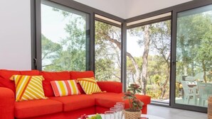 Ein leuchtend rotes Sofa mit gestreiften Kissen, ein weißer Couchtisch mit einer Obstschale und Weingläsern, und Blick auf Bäume durch große Fenster.