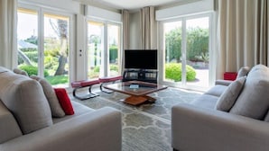 Uma sala de estar ampla com televisão de tela plana, dois sofás e uma mesa de centro. Janelas grandes proporcionam vista para o verde lá fora.