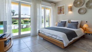 Quarto com cama, televisão e vista para uma piscina e jardim.