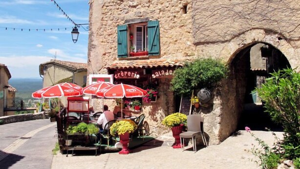 Um edifício rústico de pedra com um café chamado 'crêperie' e 'salade'. Há mesas e cadeiras ao ar livre sob guarda-sóis vermelhos.