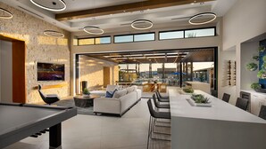 A spacious living area with a pool table, a sofa, and a dining table with chairs. There are large windows and a view of the outdoors.