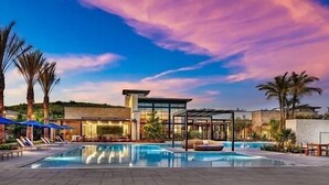 A pool area with lounge chairs and umbrellas, a modern building with glass walls, and palm trees.