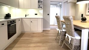 A modern kitchen with white cabinetry, a built-in oven, and a dining area with wicker chairs and a wooden table. - Tasteful vacation apartment with great attention to detail (Juist)