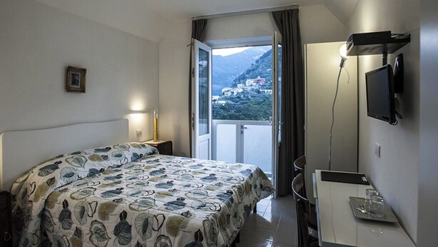 A hotel room with a bed, a desk with a computer, a TV, and a balcony with a mountain view. - Carla's House-Apartment The Olive Tree (Ravello)
