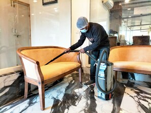A person cleaning a yellow upholstered sofa with a vacuum cleaner in a room with marble flooring and a glass door.