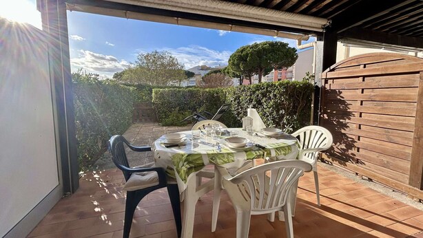 Un patio exterior con una mesa para dos, rodeado de sillas de plástico blanco, bajo una pérgola de madera.