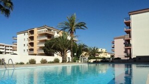 A swimming pool with clear blue water, surrounded by multi-story buildings with balconies, palm trees, and a clear sky. - Cozy 2-Room Apartment with Pool, Parking, and Near the Beach (Sainte-Maxime)