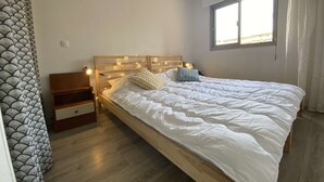 A bedroom with a wooden bed frame, white bedding, a nightstand, and a window with curtains. - Cozy 2-Room Apartment with Pool, Parking, and Near the Beach (Sainte-Maxime)