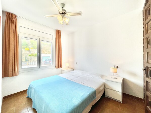 A bedroom with a bed, a nightstand, a lamp, a ceiling fan, and a window with curtains. - LES TEULES 3A Roses (Rosas / Roses)