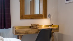 Une pièce de taille modeste, comprenant un bureau en bois, une chaise, une table de chevet, une lampe et un miroir.