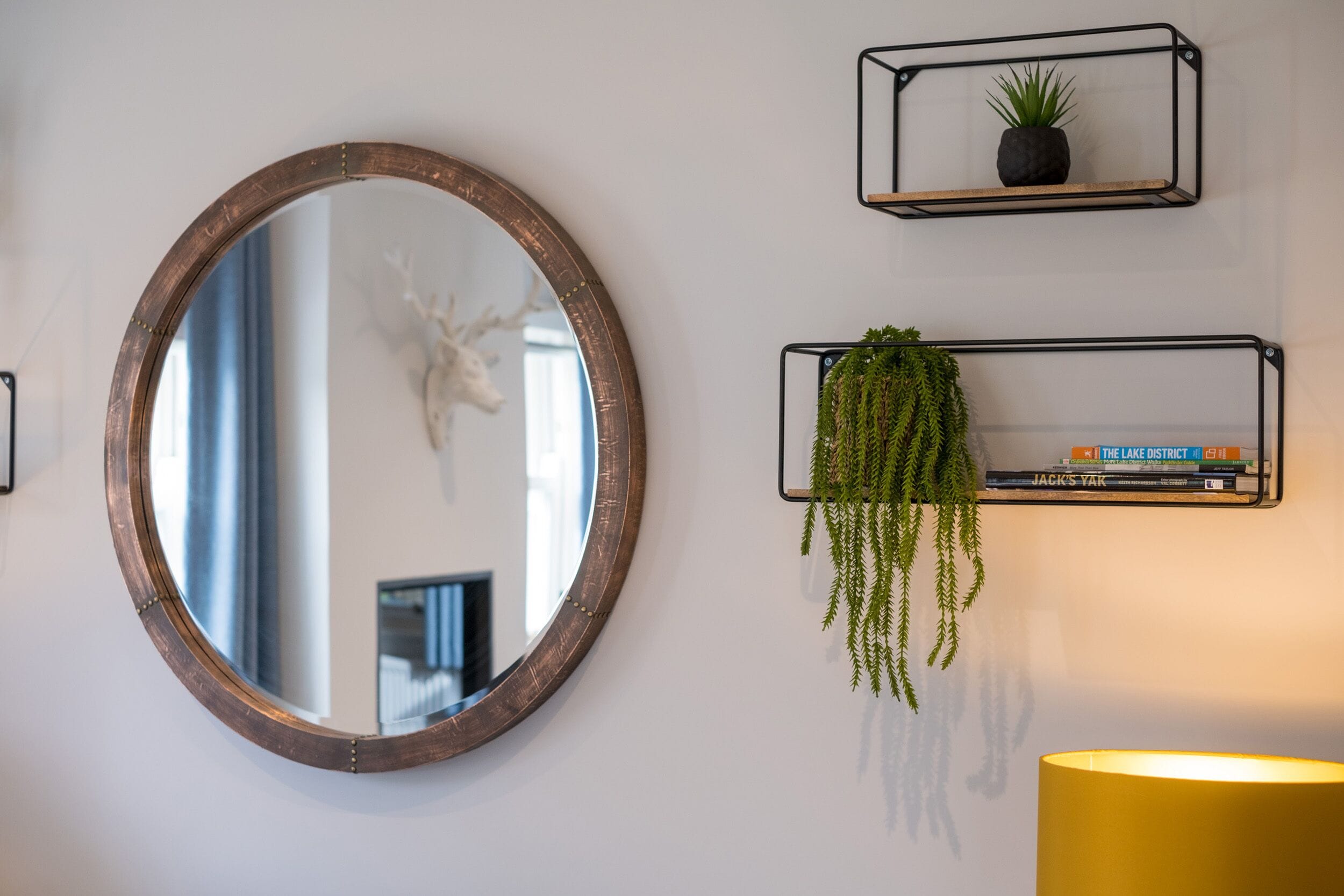 A round mirror with a wooden frame, two black metal shelves with plants and books, and a yellow lamp.
