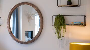 A round mirror with a wooden frame, two black metal shelves with plants and books, and a yellow lamp.