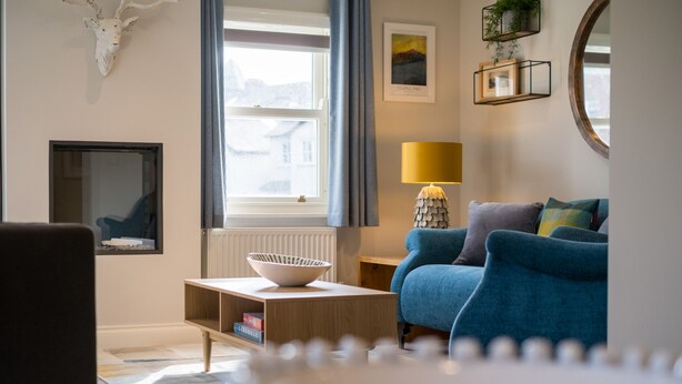 Un salón moderno con un sofá azul, una mesa de centro de madera y un cuenco blanco. Hay una ventana con cortinas, una lámpara y un cuadro en la pared.
