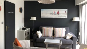 Een moderne woonkamer met een grijze bank, gestreepte kussens, een kleine witte tafel, rieten stoelen, en een groot schilderij van zeilboten aan de muur.