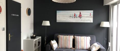 Ett modernt vardagsrum med en grå soffa, randiga kuddar, ett litet vitt bord, flätade stolar och en stor tavla med segelbåtar på väggen.