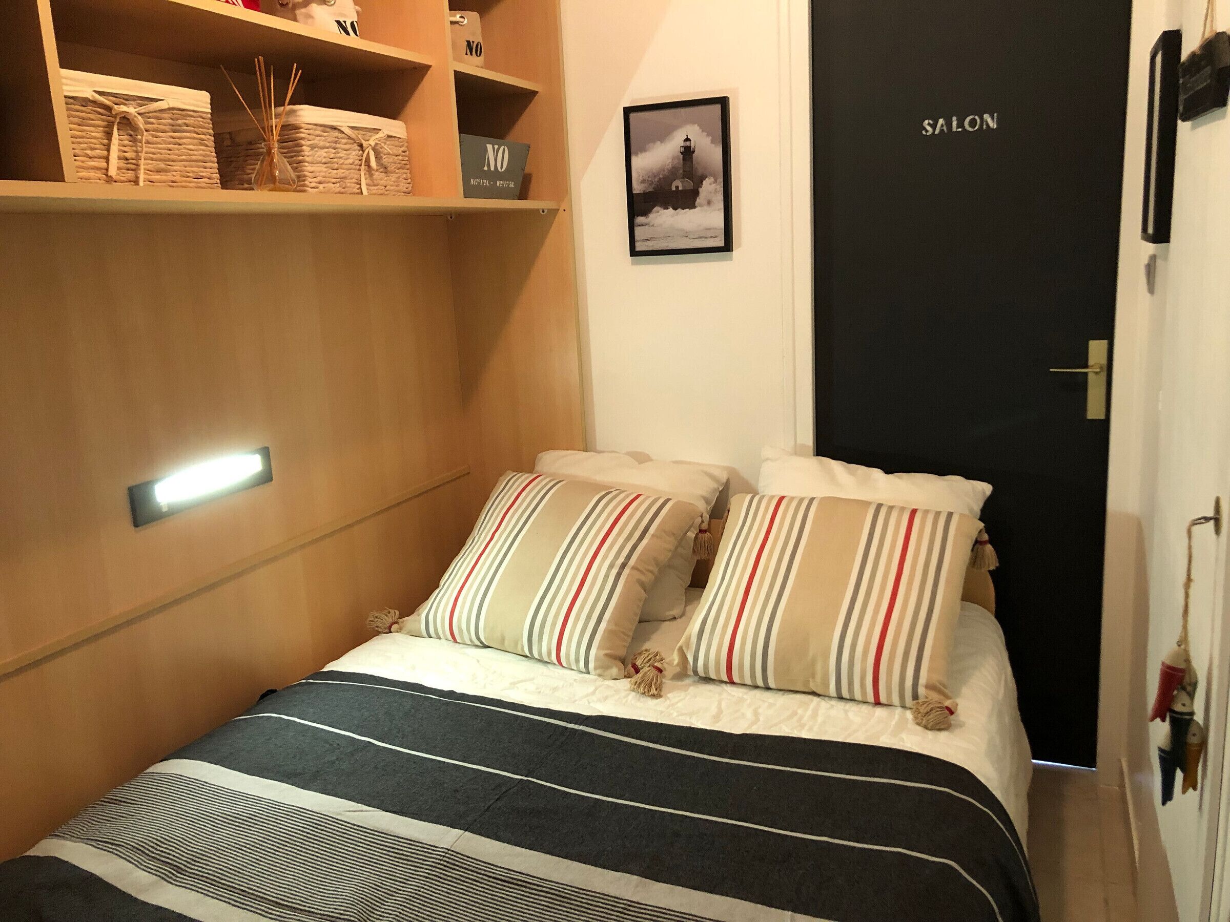 A bedroom with a bed, striped pillows, a bedside table, a wall-mounted shelf with storage boxes, a framed picture, and a door labeled "SALON".