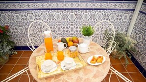 Une table ronde recouverte d’une nappe en dentelle, entourée de chaises blanches, et dressée avec des aliments pour le petit-déjeuner, notamment des fruits, des viennoiseries et des boissons.