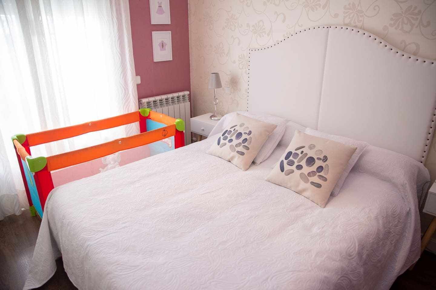 Una camera da letto con un letto bianco, due cuscini con motivo a ciottoli, un comodino con una lampada e un termosifone.