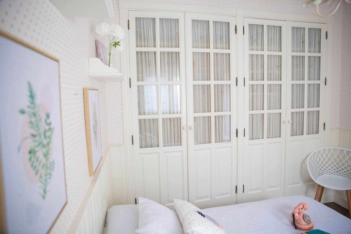 A bedroom with a bed, a white wardrobe, a chair, and a wall-mounted shelf with a plant.